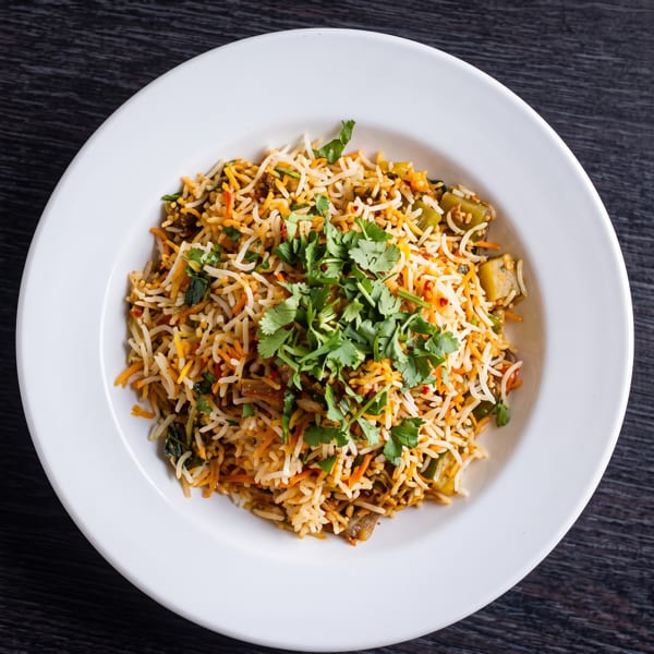A white bowl filled with colorful biryani rice, garnished with fresh green cilantro, on a dark wooden table.