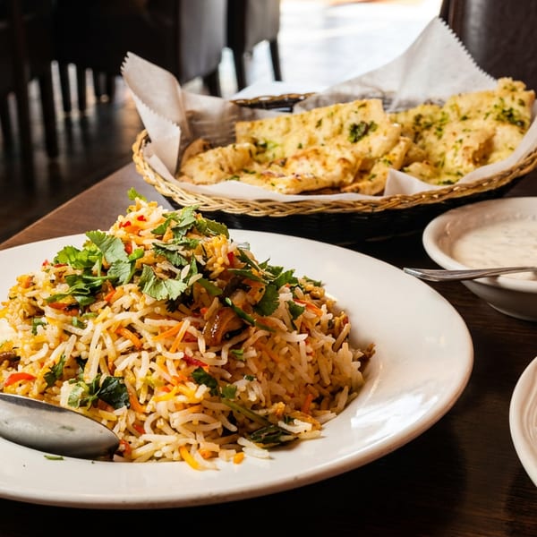 Biryani rice with cilantro, a basket of naan, and raita on a dark wooden table.